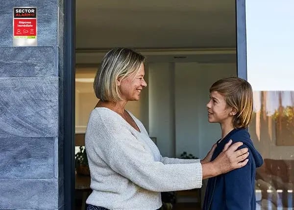Femme souriante posant ses mains sur les épaules d’un adolescent à l’entrée d’une maison.